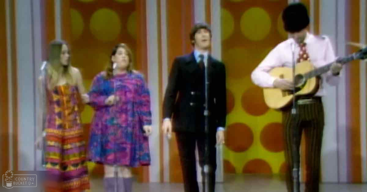 A musical performance scene from an older television show or concert setting with vibrant floral dresses for two adult females and formal attire for two adult males, suggesting a stage environment.