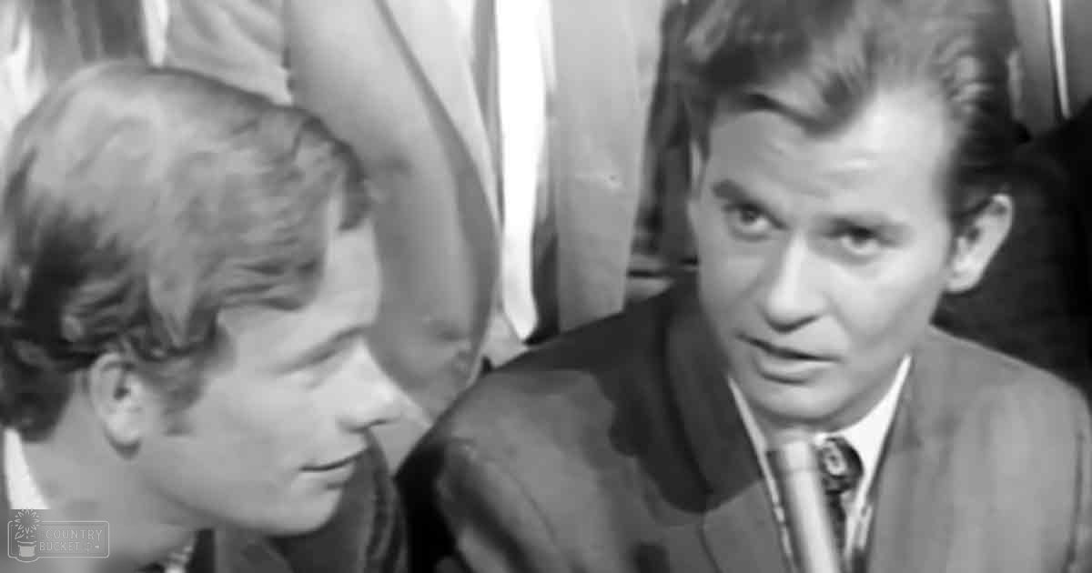 Two young adults in a mid-20th-century indoor setting with curtains visible behind them; one wearing a dark, vintage-style dress or jacket and the other dressed in formal attire.