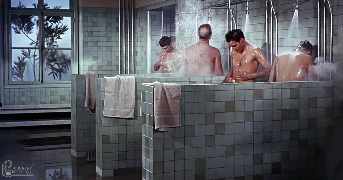 Adult males in a communal bathing area with tiled walls, hanging towels, showerheads and plants visible outside.