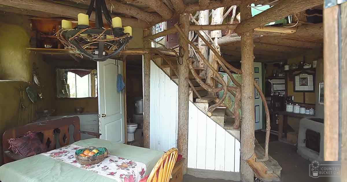A rustic cabin interior with wooden beams, natural decor, chandelier, green bedding, floral tablecloth, and kitchen items.