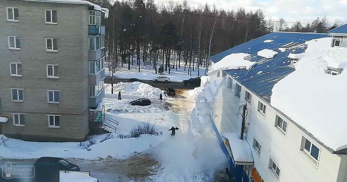 Two adult males clearing snow from a car in an urban residential area during winter, with snow-covered trees and buildings surrounding the scene.