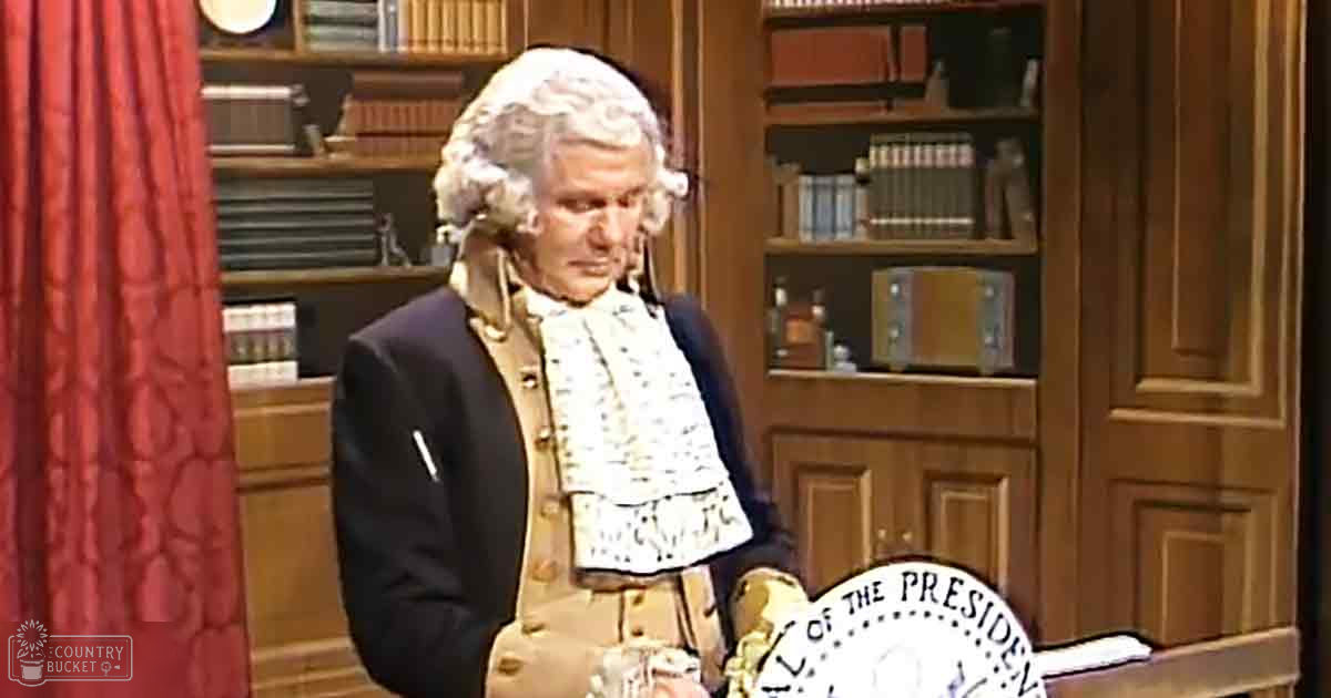 An older adult male in an 18th-century-style black coat with gold buttons and white cravat, seated amidst wooden furniture filled with books in a library or study room setting.