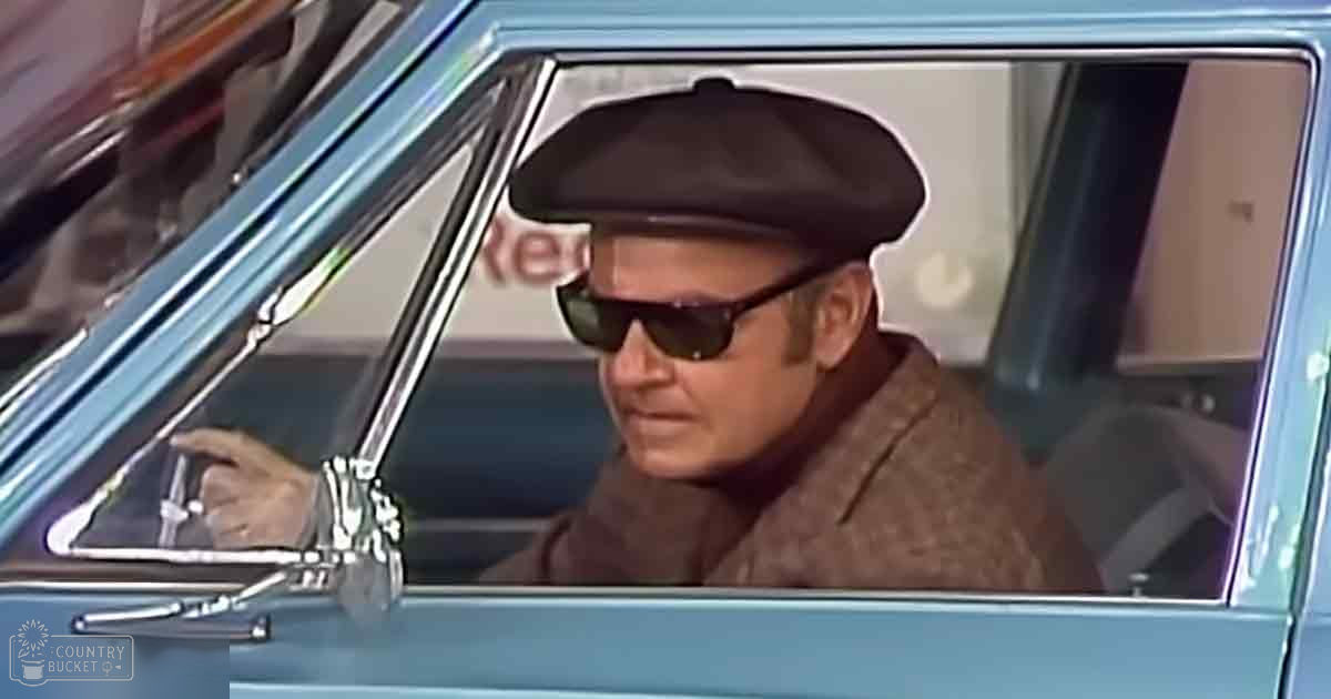 An older adult male wearing a brown beret-style hat and dark sunglasses, dressed in what appears to be a mid-20th century style brown coat or jacket, sitting inside an older model car with chrome detailing on the dashboard and side mirror. The setting is outdoors during daytime.