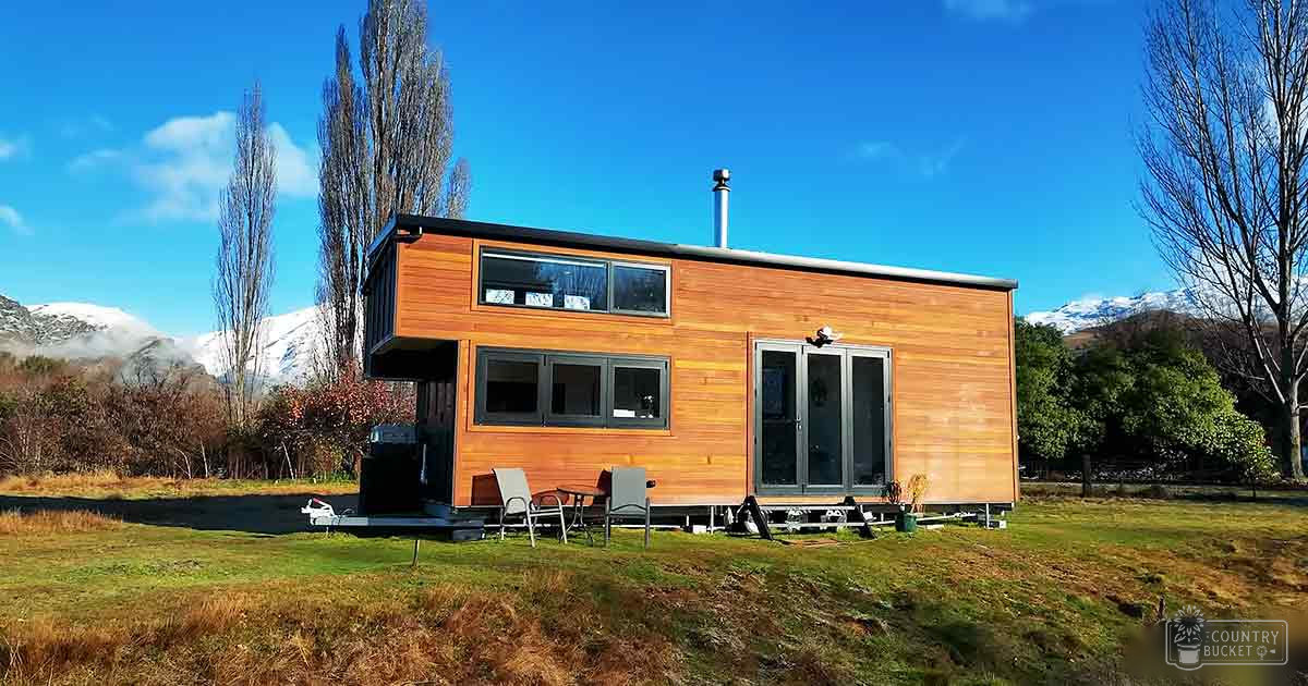 A serene rural scene with a modern wooden cabin surrounded by snow-capped mountains under clear blue skies.