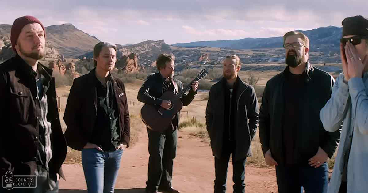 Group of adult male musicians with acoustic guitar, red rock desert landscape, partly cloudy sky at late afternoon or early evening.