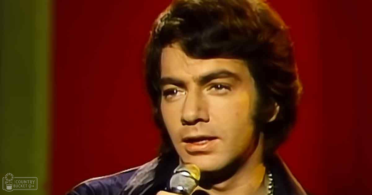 A young adult male in a textured blazer from the 1970s era performs at an event or concert against a vibrant red and green backdrop.