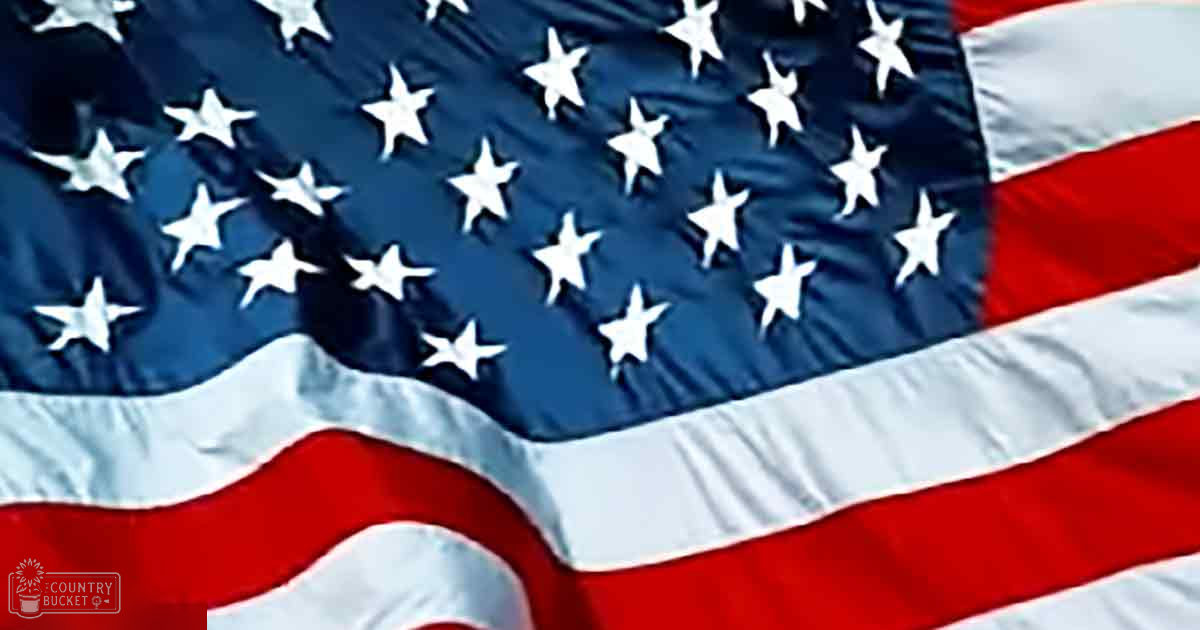 American flag with white stars on a blue field and red and white stripes, set against an outdoor backdrop during daylight hours.