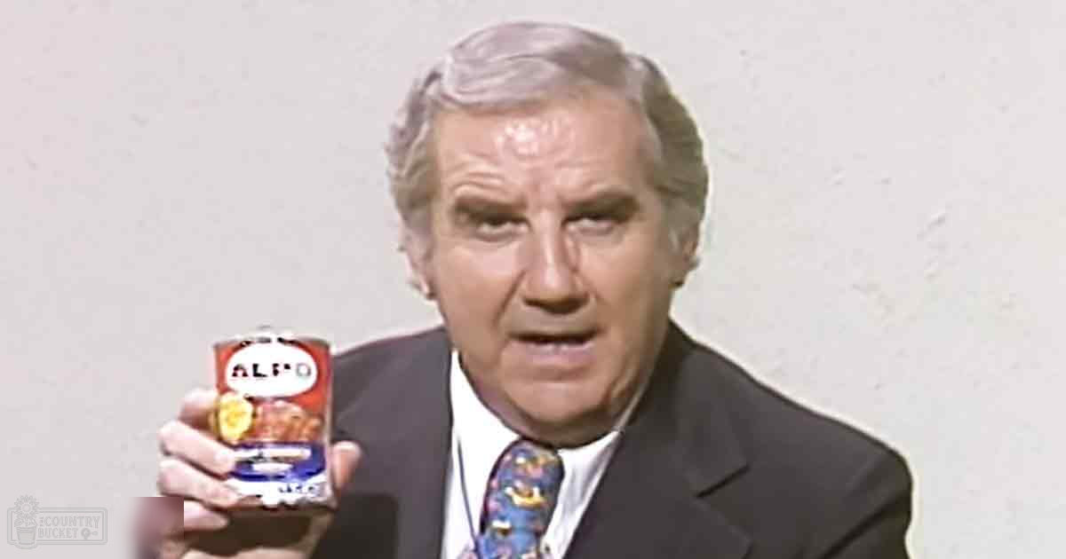 A middle-aged adult male in a formal dark suit with a white dress shirt and colorful tie, holding a can labeled 'ALF-O' against an indoor off-white background.