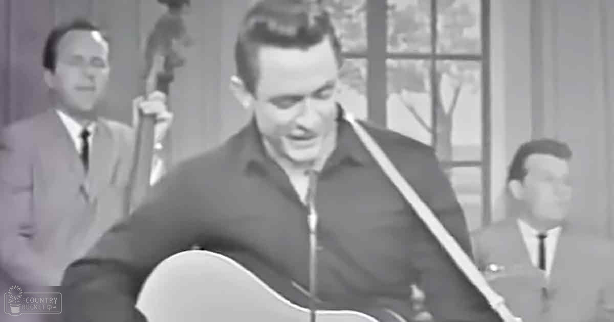 A man playing an acoustic guitar in a studio setting with two background individuals wearing mid-20th century suits; dimly lit, possibly daytime through the window.