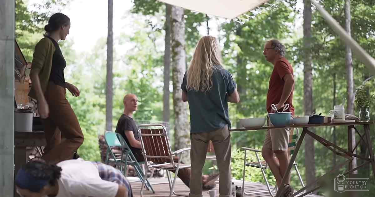 Adults gathered outdoors in a rustic campsite setting with folding chairs and simple meal preparation items.