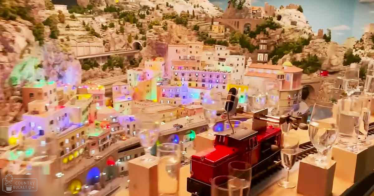 A detailed model town with miniature buildings, a red train on tracks, and wine glasses in an indoor setting resembling a museum exhibit.
