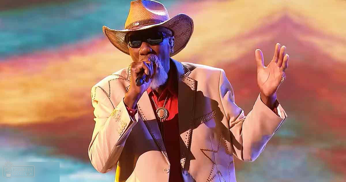 An older male singer wearing a beige jacket with intricate embroidery, red shirt, dark vest or undershirt, sunglasses, and a wide-brimmed hat, performing on stage with a microphone. The vibrant background suggests an evening performance setting.