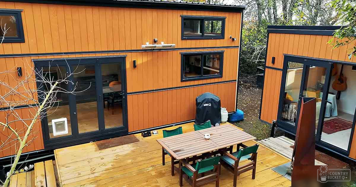 A residential outdoor setting with a tiny house, wooden dining area, grill, propane tank, guitar inside, and household decor; spring or summer day with greenery in the background.