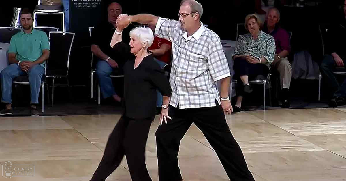 A couple dancing in a lively indoor dance studio or event space, surrounded by spectators and chairs.