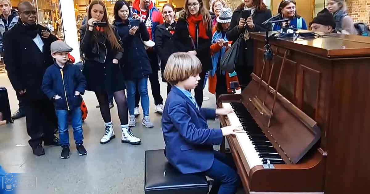 A young boy wearing a blue blazer plays piano in an indoor public space with several adults and microphones nearby.