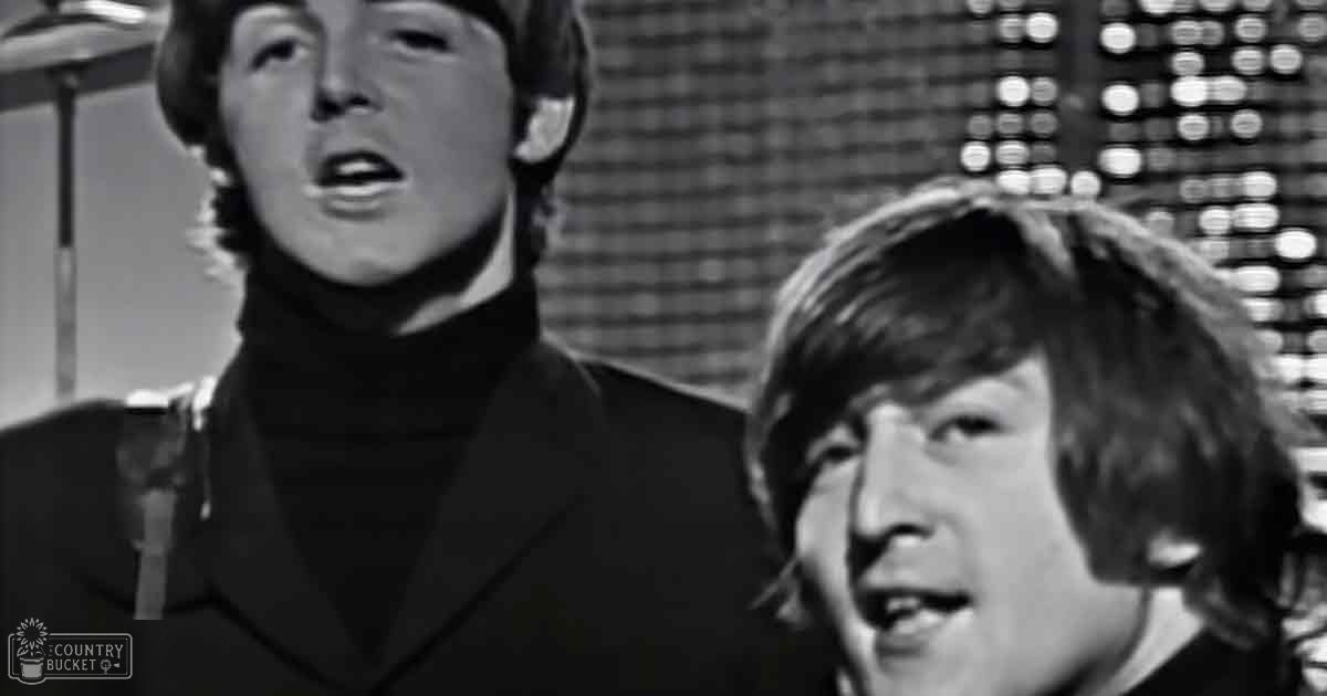 Two young adult males in a mid-20th century performance setting, wearing dark-colored turtleneck sweaters and suits, with stage lighting and a drum set visible in the background.
