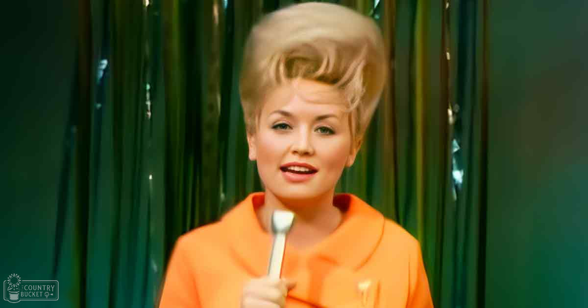 A middle-aged woman wearing a bright orange vintage-style garment with 'Madly Odd' text, holding a spoon, against green curtains indoors.