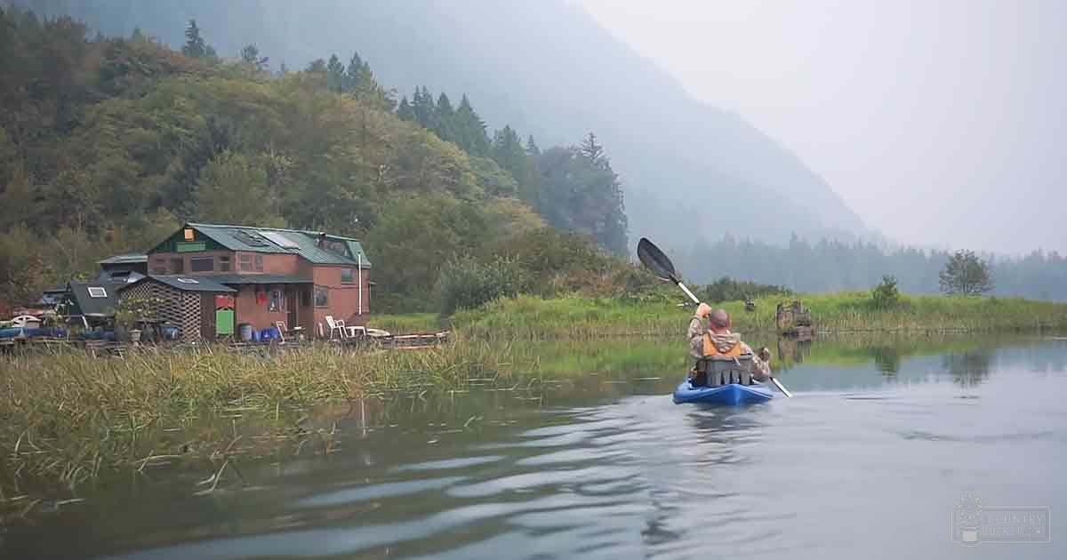 A middle-aged male kayaking near a lakeside cabin amidst lush greenery with mountains shrouded in mist, under an overcast sky.