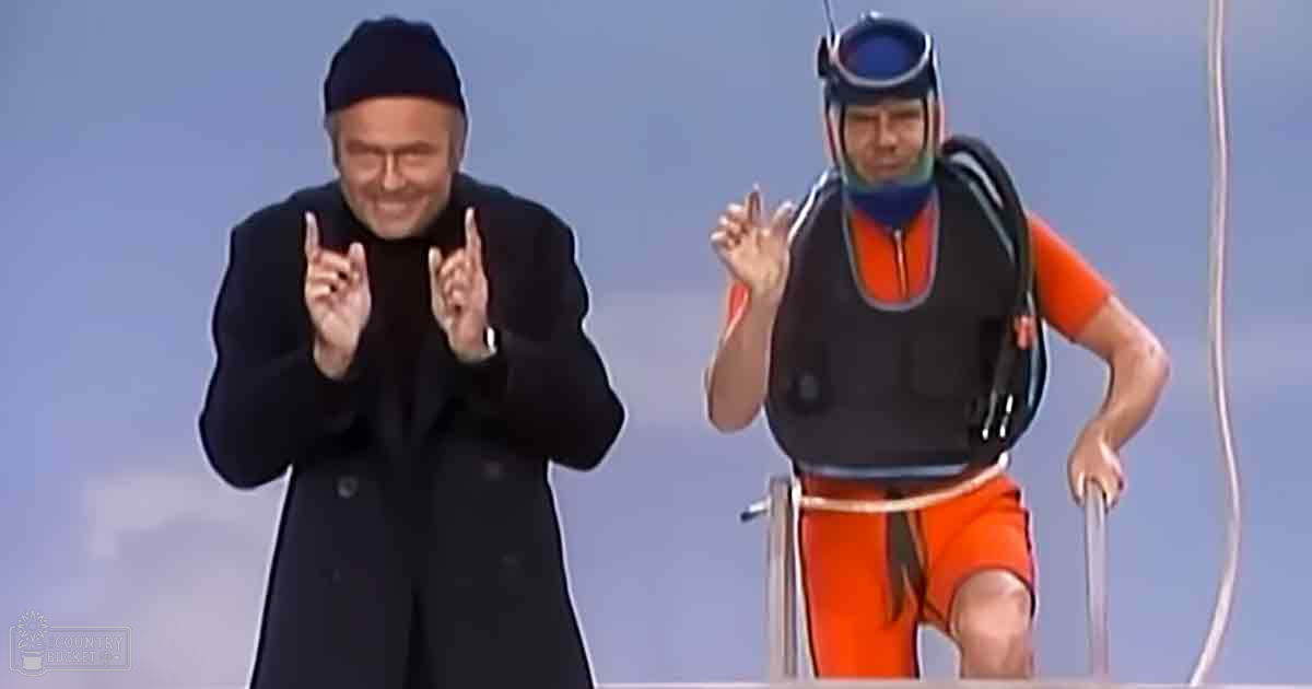 An adult male dressed in a black coat and pants stands beside another adult male in an orange swimsuit with a life vest, indicating a comparison or interaction between two individuals in contrasting attire within an indoor setting.
