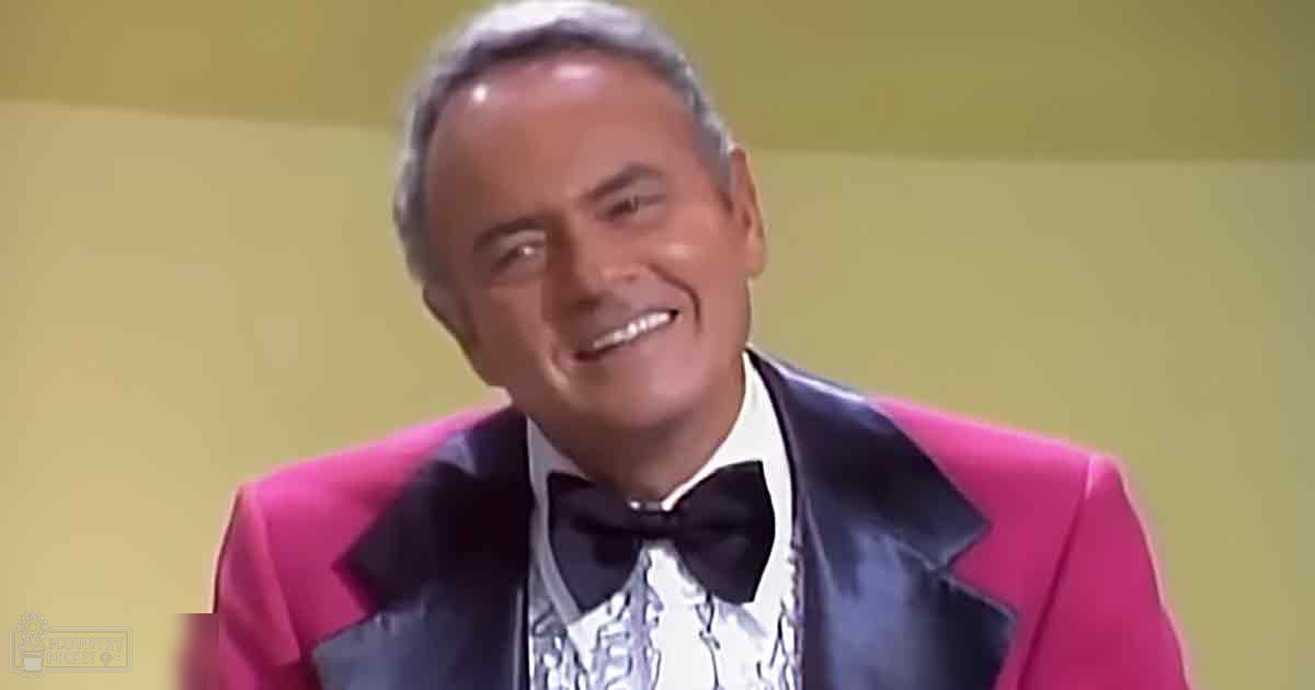 An older adult male in a formal setting wearing a bright pink tuxedo jacket with black lapels and a white patterned shirt under it, possibly from the mid-20th century.