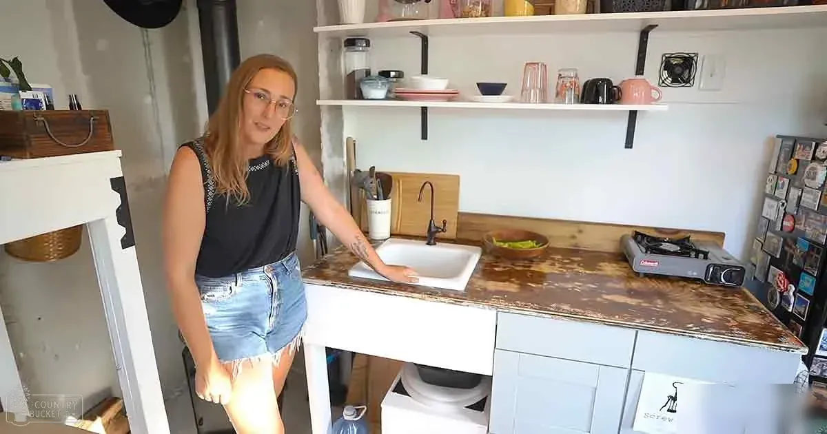 A young adult female stands in a well-lit kitchen, wearing a black sleeveless top with decorative details at the neckline and light blue denim shorts. She is near open shelving holding various items such as cups, bowls, and a blender.