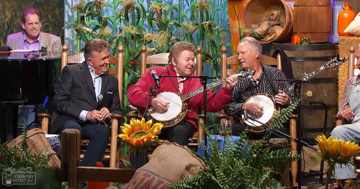 A musical performance set in an indoor barn-themed studio with greenery and rustic decor with a piano, banjos, microphones, sunflowers, and barrel-like structures.