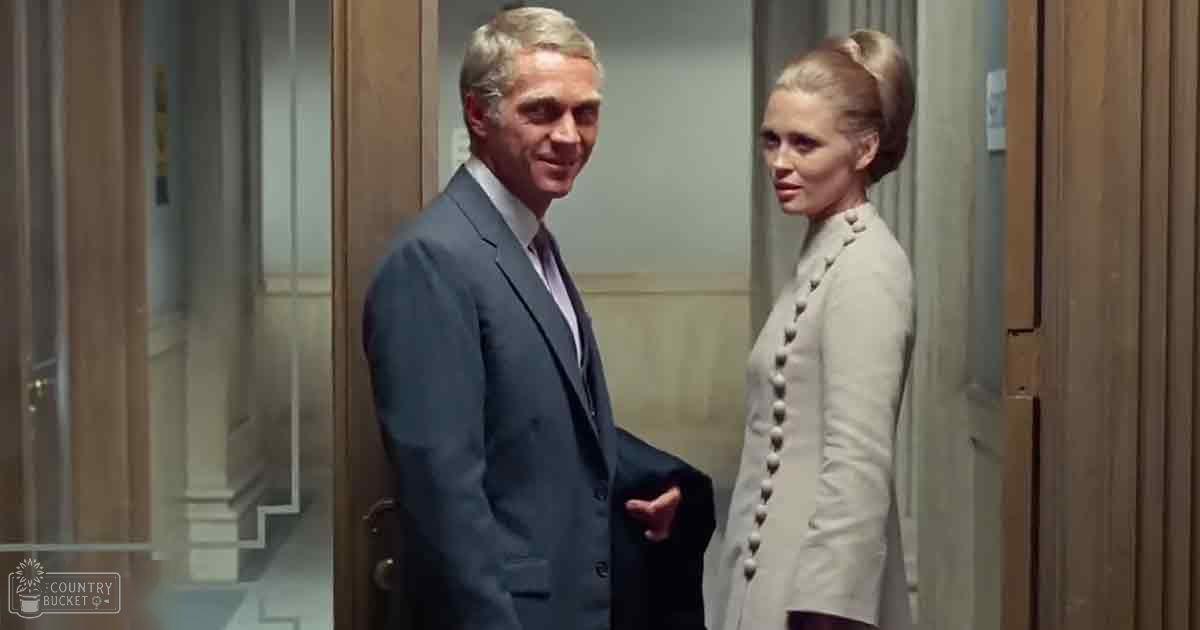 Adult male singer in a dark gray suit with light purple shirt, middle-aged female wearing a structured light gray coat and updo, formal indoor hallway with wooden paneling, even bright lighting suggesting daytime.