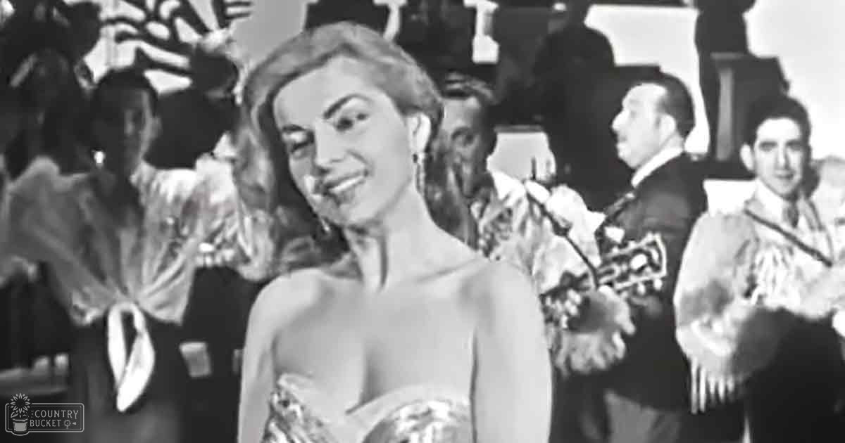 An adult female in a formal gathering wearing an intricate strapless gown with embroidery, likely from the mid-20th century.