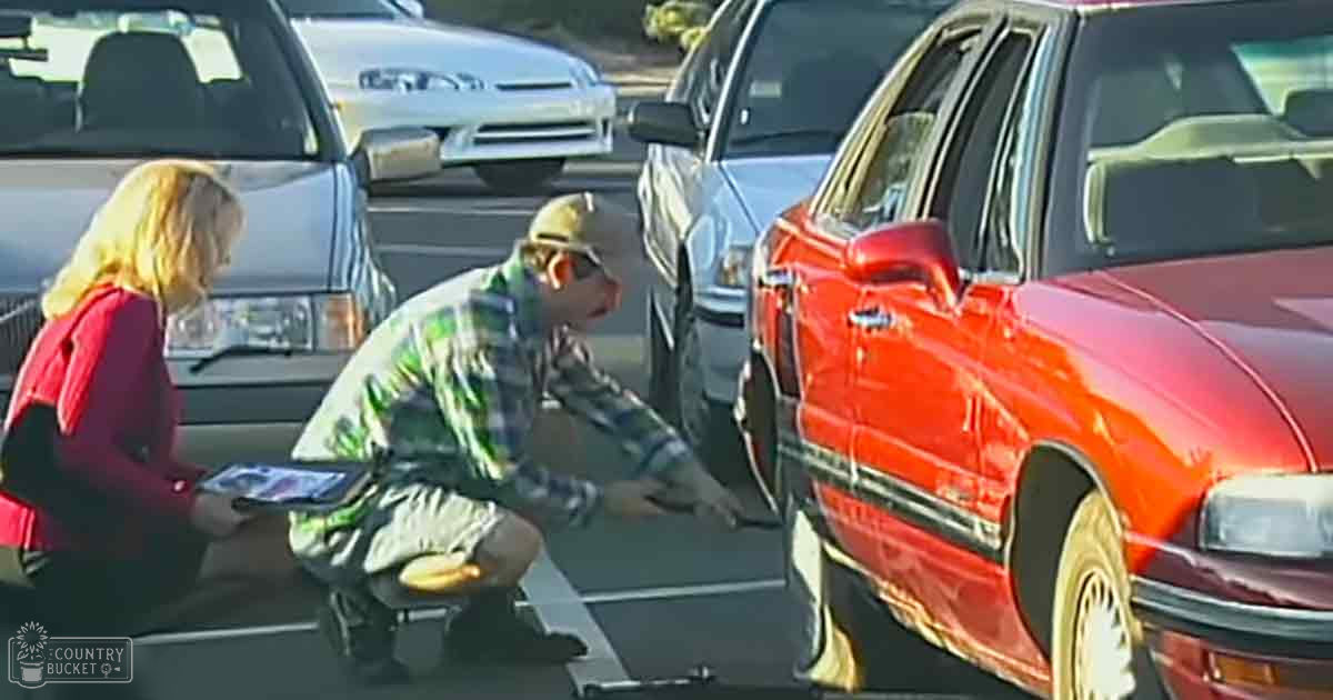 Adult male wearing a plaid green and white shirt, beige cap, and sunglasses works on a red sedan in an outdoor parking lot during daylight with clear weather conditions.