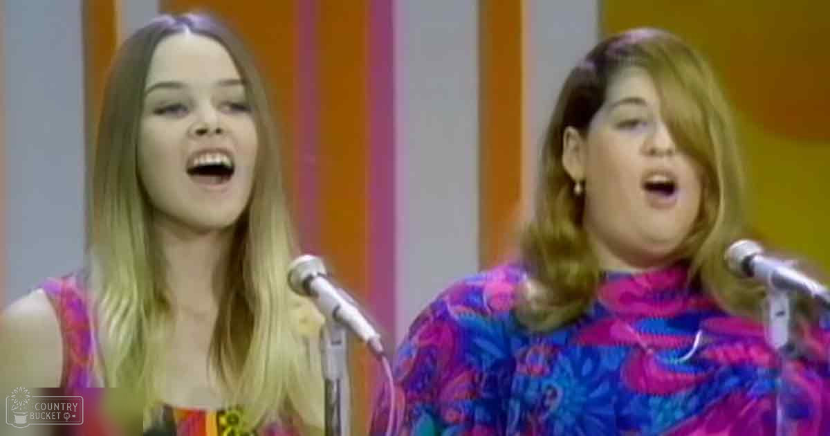 Two young adult females singing in a studio setting, wearing sleeveless floral tops and patterned dresses from the 1960s or early 1970s with microphones on stands.