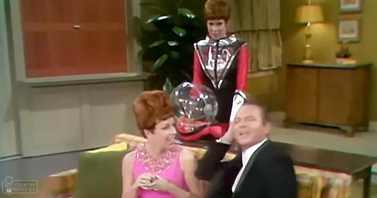 A mid-20th century living room scene with an adult female in a pink dress, seated male in formal attire, and performer in a metallic outfit; green armchair, wooden sideboard with lamp and framed pictures, potted plant visible.