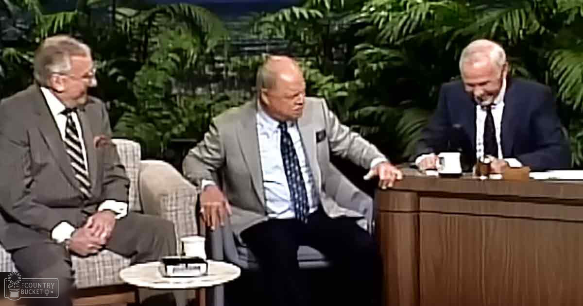 Three men in a talk show set with formal attire; dark gray suit jackets, plaid shirts, indoor studio setup designed to look like a natural environment.