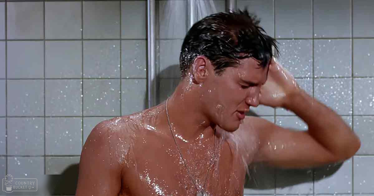 A young adult male stands shirtless in a tiled shower, wearing a necklace; bright daylight illuminates the scene.