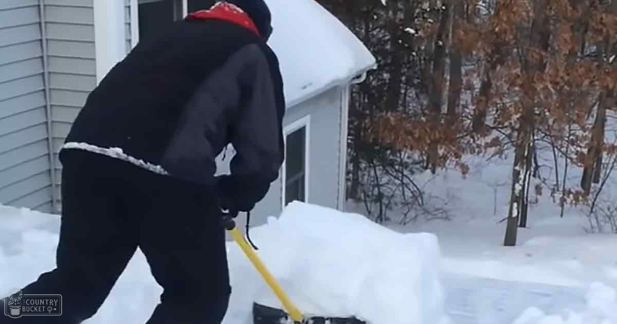An adult male shoveling snow outside a house with bare trees indicating winter, under natural daylight.