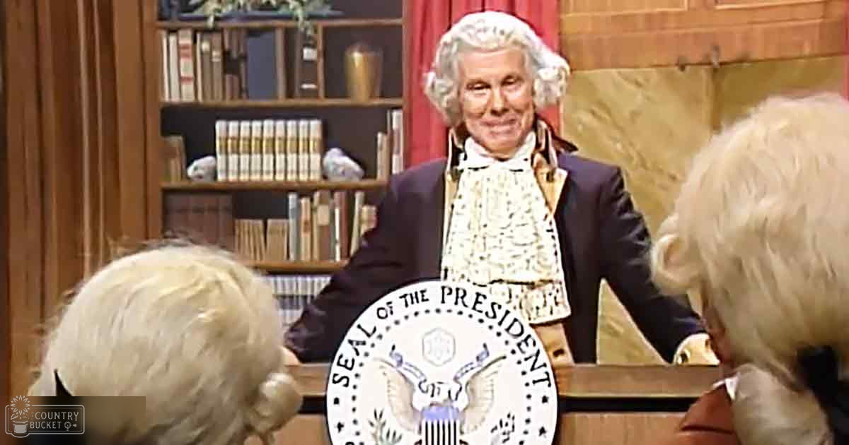 An older adult male impersonating George Washington in front of a historical reenactment audience dressed in period attire. The setting is an indoor library or study room with wooden furniture, red curtains, and bookshelves filled with books.