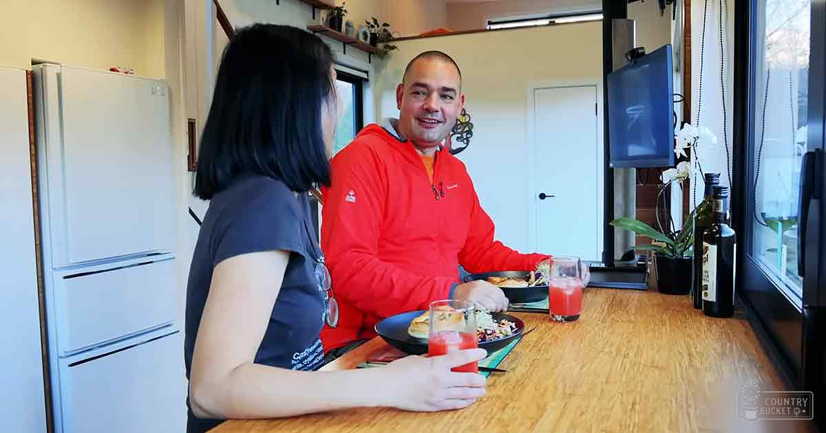Two adults enjoy a meal together in their home kitchen with natural light streaming through windows, surrounded by houseplants and modern decor.