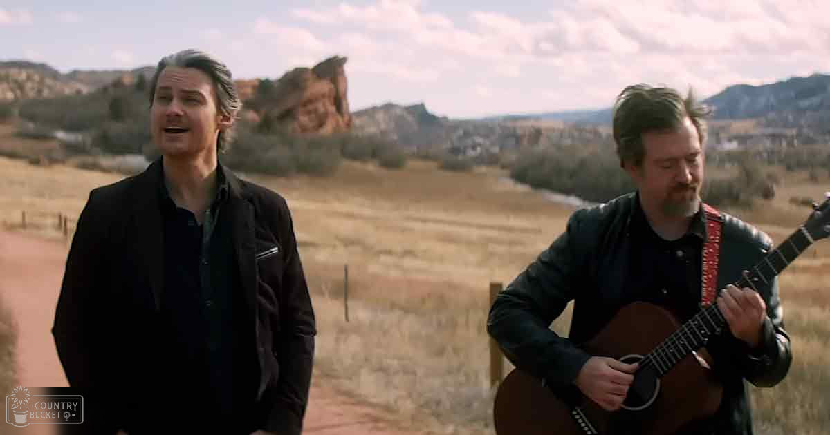 Two adult males performing music outdoors on a dirt path with red rock formations and partly cloudy sky, late afternoon or early evening.