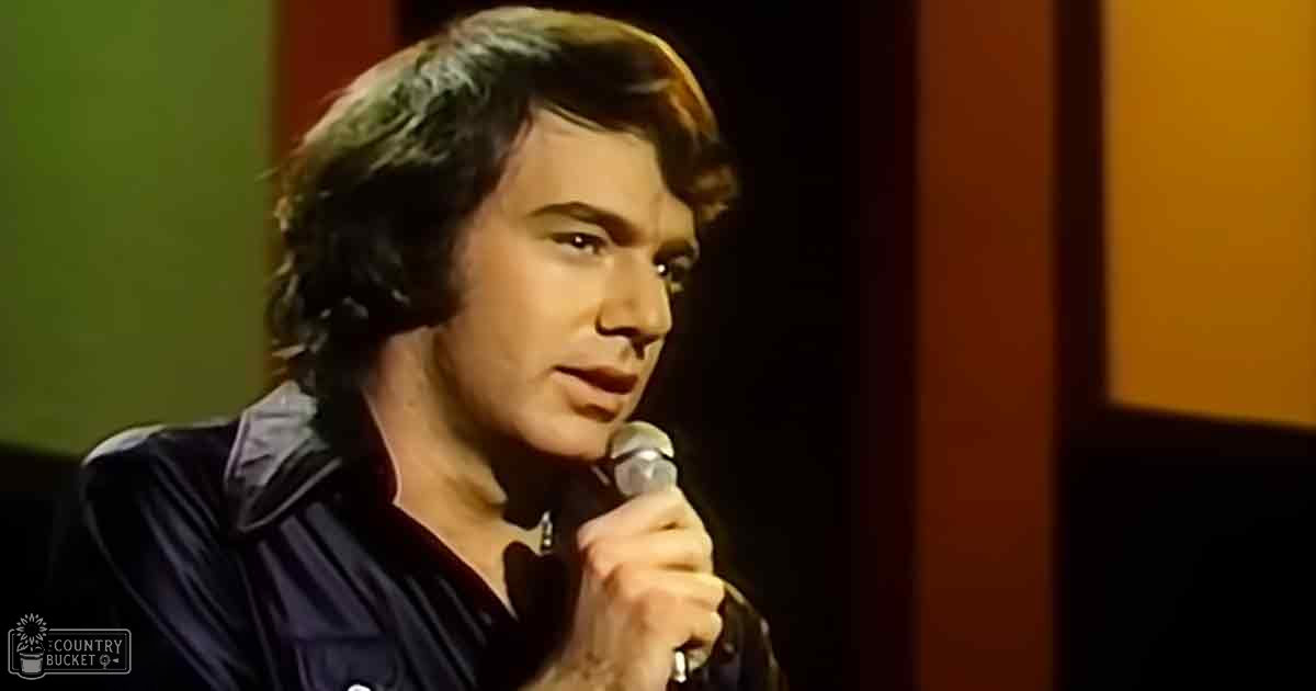 A young adult male singer wearing a long-sleeved dark blue button-down shirt, performing on stage with a microphone held by him against a dark curtain backdrop under artificial lighting.