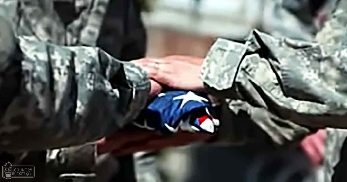 A military ceremony with two adults in camouflage uniforms holding a small American flag during daytime outdoors, with natural light and blurred greenery in the background.