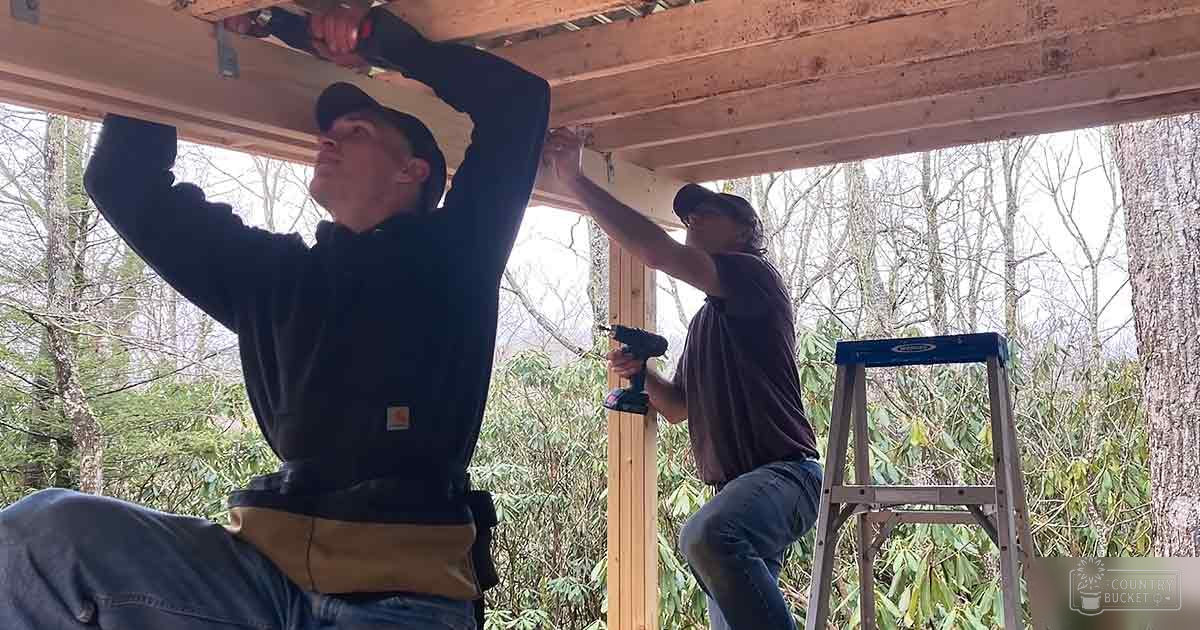 Two adults working together outdoors with construction equipment and a blue step ladder amidst trees suggesting spring or summer.