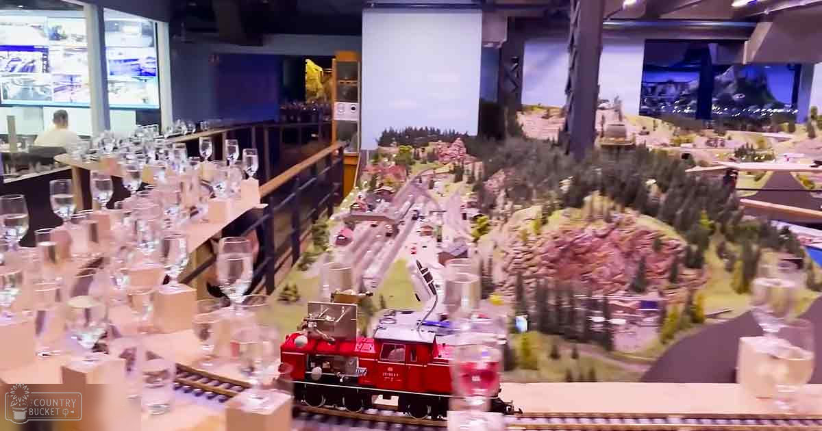 Model train setup with miniature landscapes, wine glasses, and champagne flutes in an indoor exhibition space.