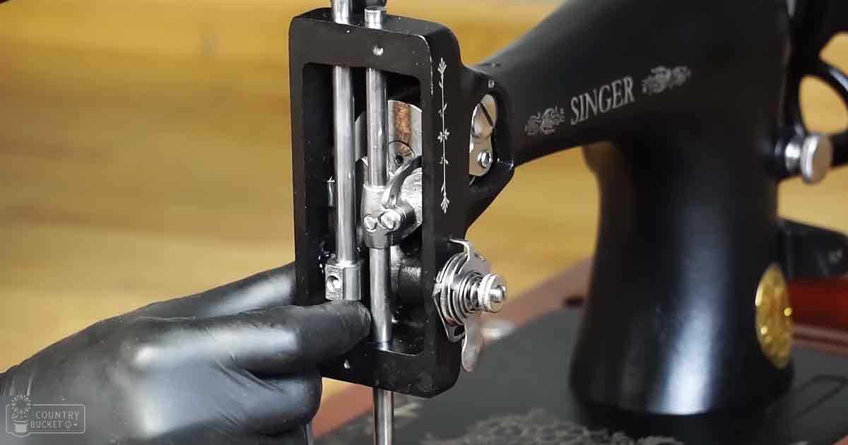 A close-up view of an older Singer sewing machine's needle clamp mechanism, indoors on a wooden workbench with natural or artificial lighting.