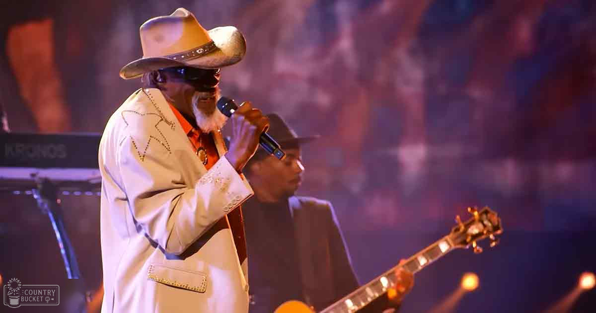 An older adult male performer in a light-colored suit jacket with decorative stitching, red shirt, and wide-brimmed hat on stage at an indoor concert venue.
