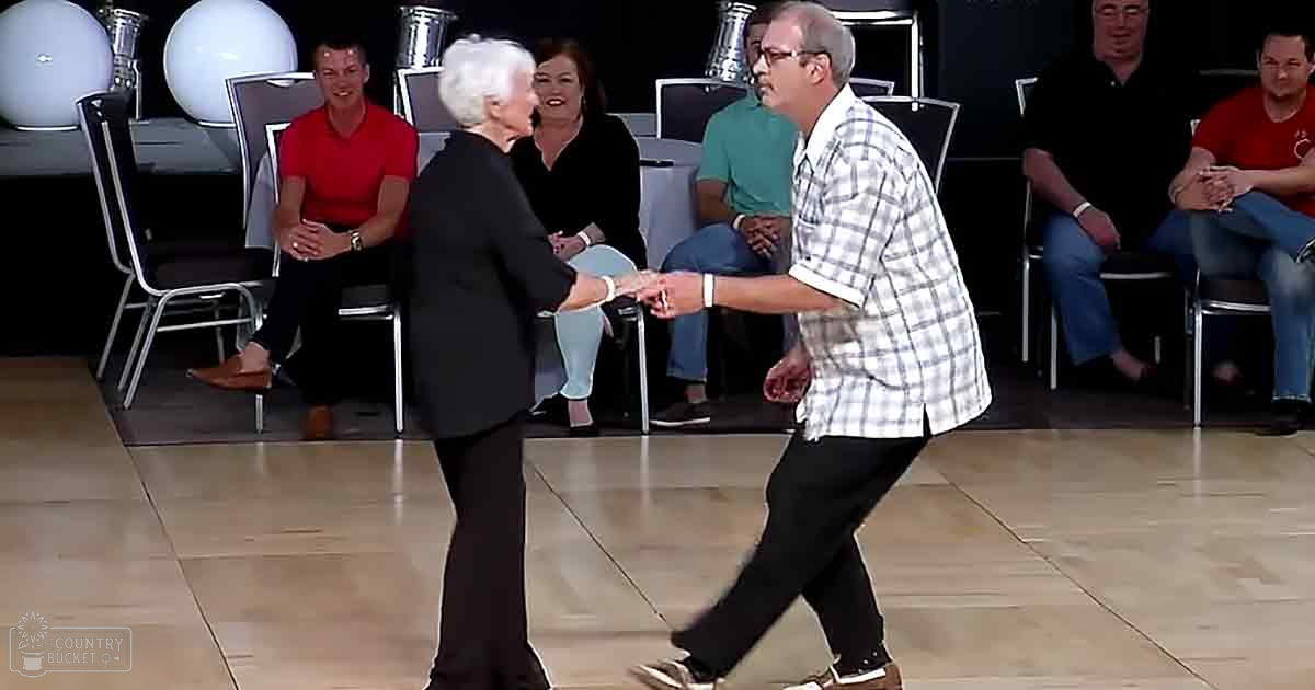 An older adult female with short white hair in a black blazer stands among other adults in an indoor dance scene; modern chairs and spherical light fixtures are part of the setting