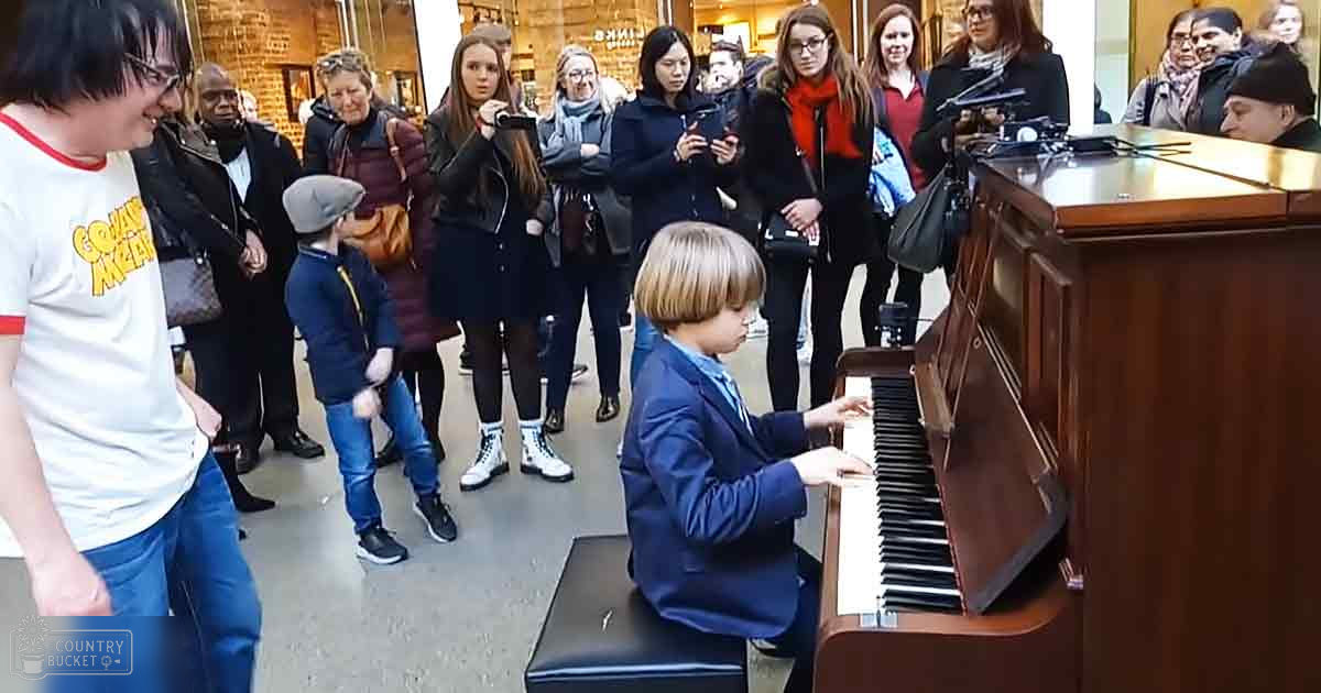 A young child in a formal uniform plays the piano indoors, surrounded by adults and casual observers; brick wall and display case in background suggest museum or gallery setting.