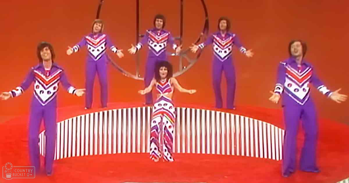 A performance group with four adult males in matching purple tracksuits and a female performer wearing a colorful, patterned outfit in a studio setting. The backdrop is orange, suggesting an indoor stage setup.