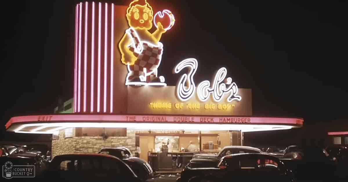 A parking lot at Bob's Big Boy restaurant at night with illuminated neon sign in pink and yellow, vehicles from mid-20th century era parked nearby under dark skies.