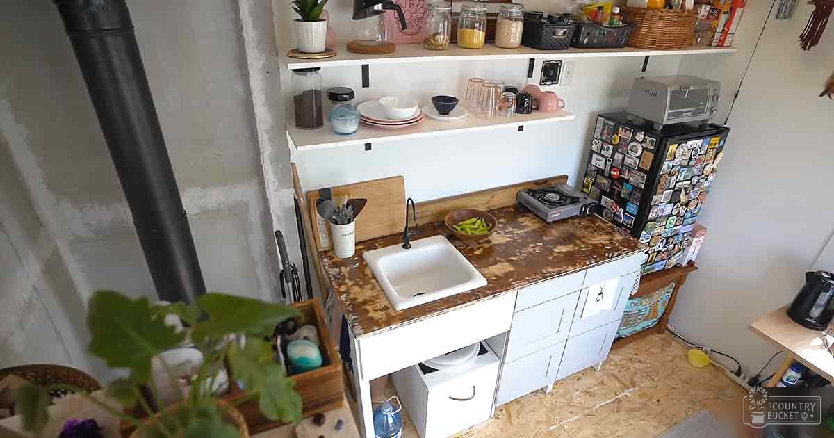 Compact kitchen setup with a white sink, brown speckled countertop, black refrigerator adorned with stickers, portable gas stove, and potted plant in an indoor space.