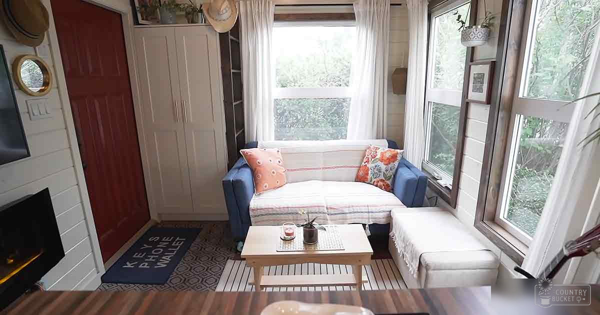 A cozy living space with a blue sofa, wooden coffee table, white curtains, and geometric-patterned rug in a small cabin or home setting.