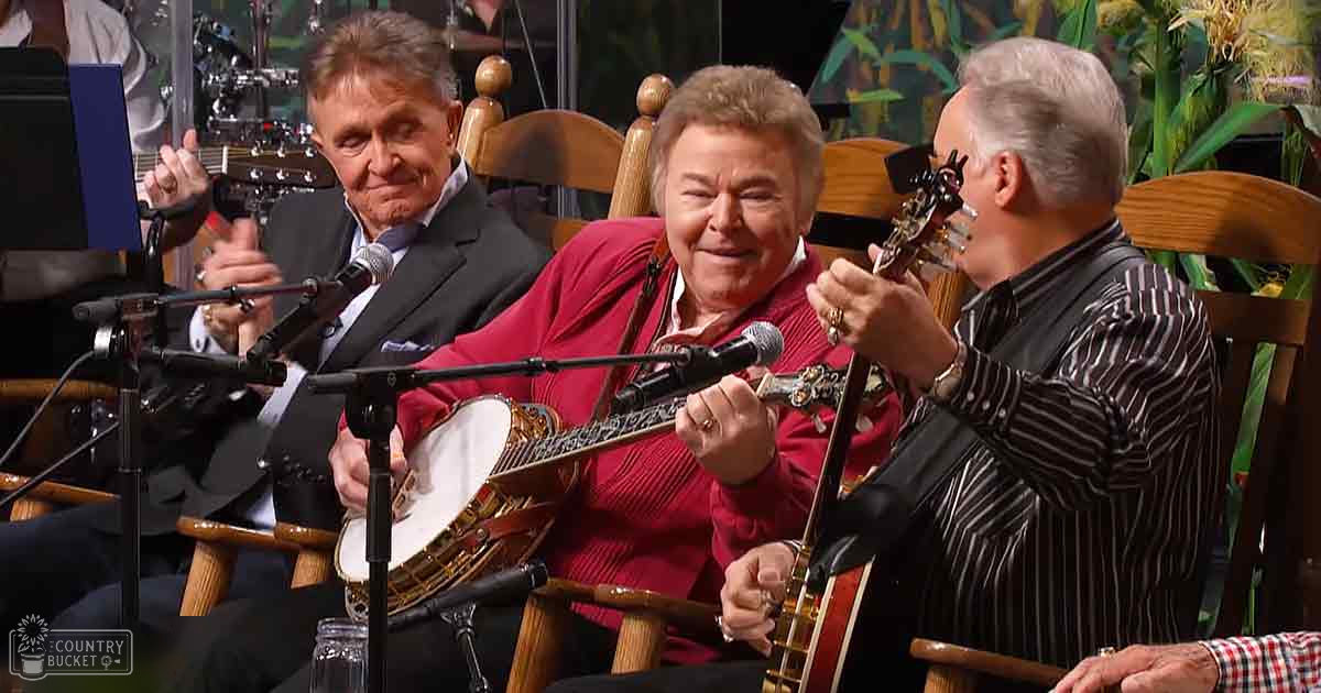An older adult male singer in a dark suit with a white shirt and tie, holding a guitar; another older adult male in a red sweater vest over a collared shirt, possibly button-down or dress shirt, with a microphone; a third older adult male in a black and white striped long-sleeve shirt with collar, wearing a watch on the left wrist. Indoors, likely a stage or studio setup for a musical performance.